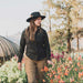 Woman in a black jacket and hat standing among flowers with trees in the background