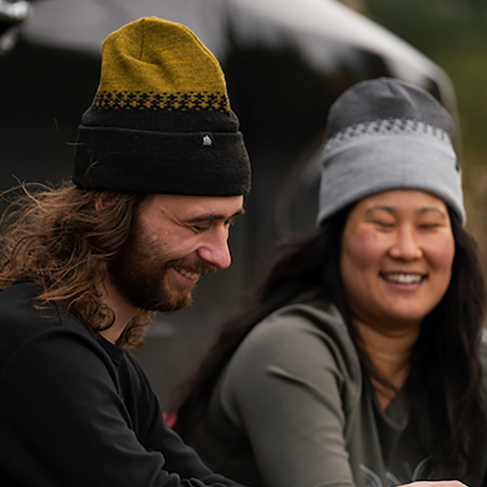 Two people sitting outdoors, one wearing a yellow and black beanie, the other a gray beanie.