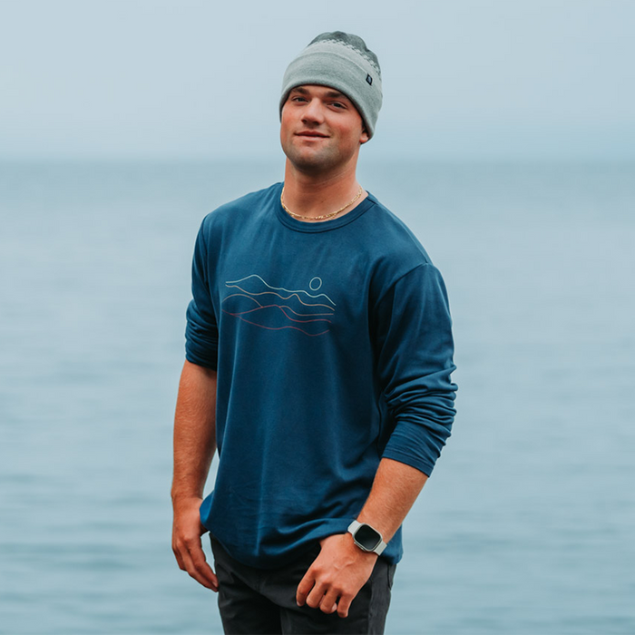 Man wearing a blue long-sleeve shirt and gray beanie standing in front of a body of water.
