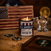 Candle in a container labeled 'All-American Pure Motor Oil' on a wooden surface with an American flag in the background.