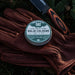 Solid cologne tin on brown leather gloves with a knife in the background