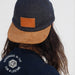 Person wearing a navy cap with brown brim and leather patch, back view.