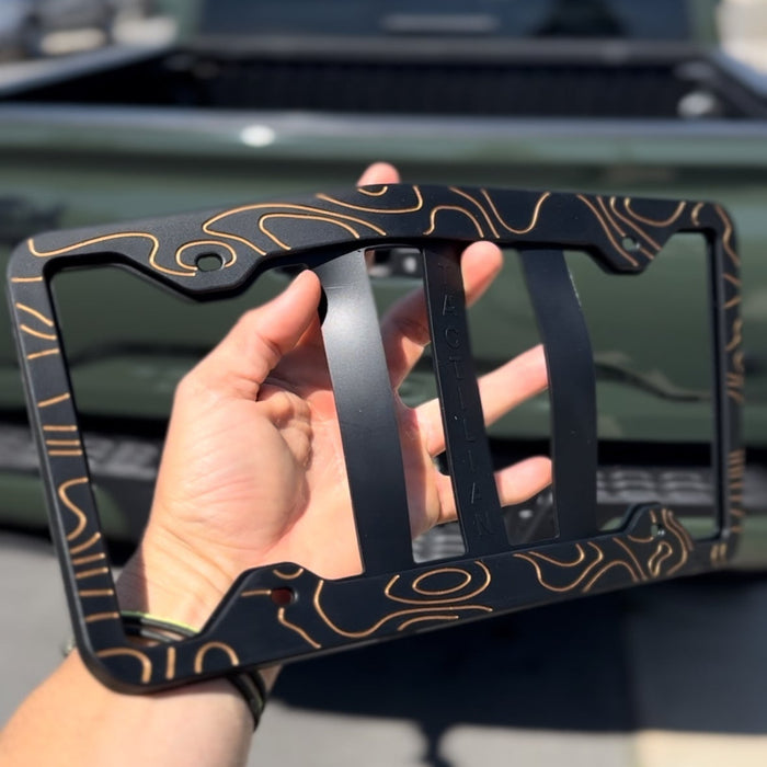 Person holding a decorative black metal license plate frame with a blurred green truck in the background.