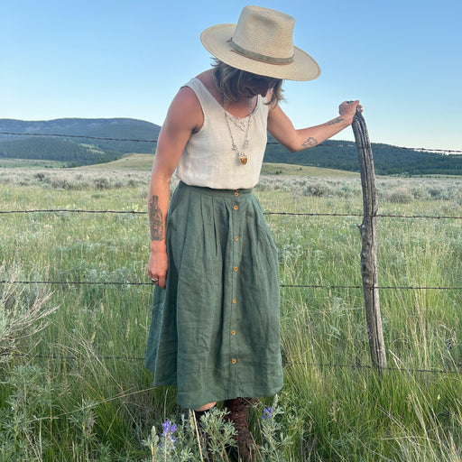 Woman in a green skirt and beige hat standing in a field with mountains in the background