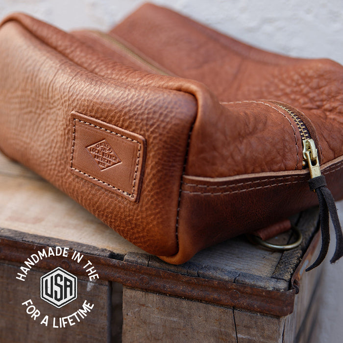 Brown leather toiletry bag with a visible brand logo on a wooden surface.