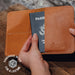 Brown leather passport holder with a passport inside, held by a hand on a wooden surface.