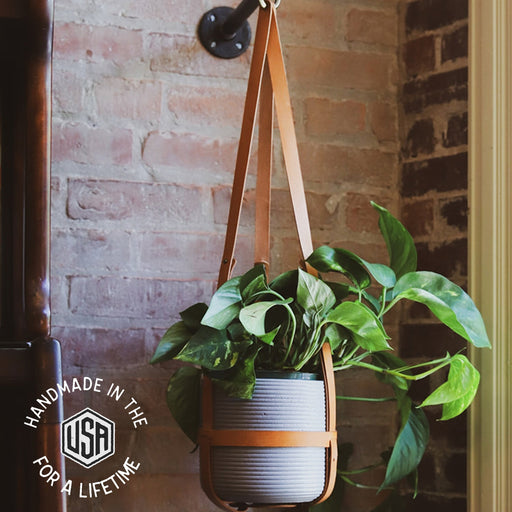 Hanging plant holder with leather strap against a brick wall, featuring 'Handmade in the USA for a Lifetime' logo.