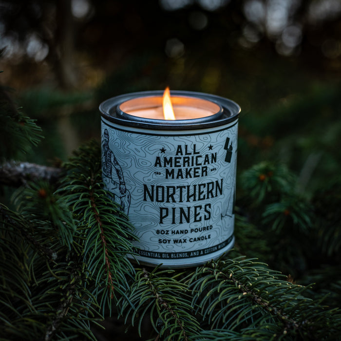 Candle in a container labeled 'All American Maker Northern Pines' on a branch with a dark background