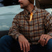 Man sitting by a fire outdoors, wearing a plaid shirt and beanie.