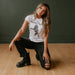 Woman wearing a white t-shirt with a graphic design, sitting on a wooden floor against a dark green wall.