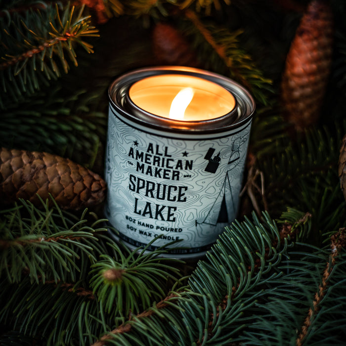 Candle in a tin labeled 'All American Maker Spruce Lake' surrounded by pine branches.