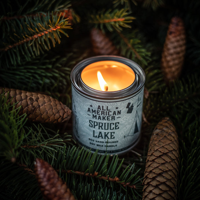 Candle in a container labeled 'American Maker Spruce Lake' surrounded by pine branches and cones.