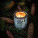 Candle in a container labeled 'American Maker Spruce Lake' surrounded by pine branches and cones.