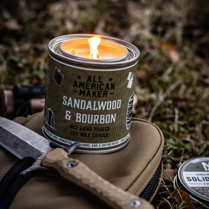 Candle in a can labeled 'All American Maker Sandalwood & Bourbon' on grass with a knife and other items.