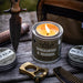 Candle in a tin labeled 'Old American Maker Sandalwood & Bourbon' with a knife, leather bag, and other items on a wooden surface.
