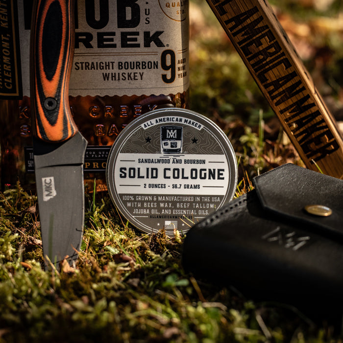 Bottle of OB Reek bourbon, knife, and Solid Cologne on a grassy surface