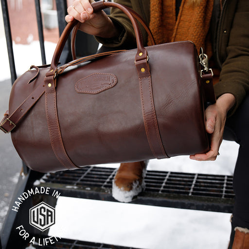 Brown leather duffel bag held by a person on a snowy street, with 'Handmade in USA for a Lifetime' label.