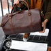 Brown leather duffel bag held by a person on a snowy street, with 'Handmade in USA for a Lifetime' label.