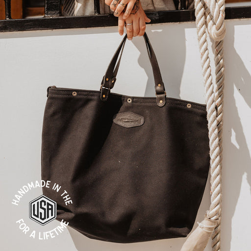 Black handbag held by a person with tattoos, hanging on a white wall.
