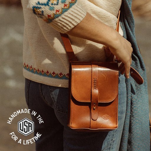 Person wearing a beige sweater with colorful patterns, holding a brown leather bag against a blurred natural background.