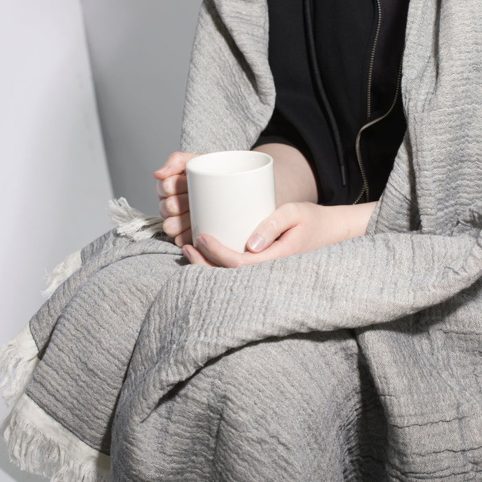 Person holding a white mug wrapped in a gray blanket against a neutral background