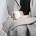 Person holding a white mug wrapped in a gray blanket against a neutral background