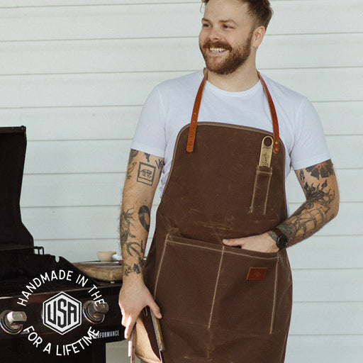 Man wearing a brown apron with a white shirt, standing against a light wooden panel background.