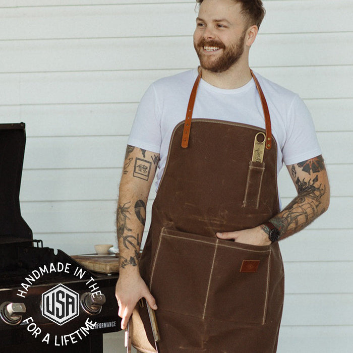Man wearing a brown apron with a white shirt, standing against a light wooden panel background.